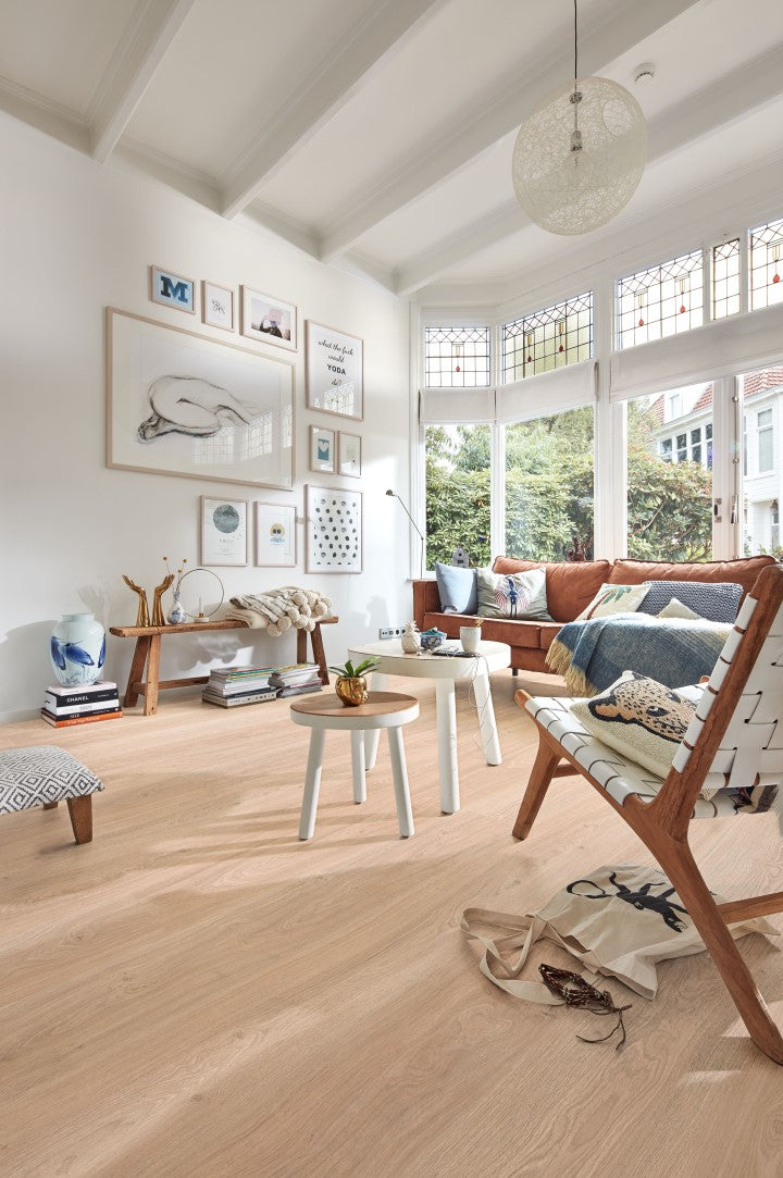 Helles Wohnzimmer mit großem Fenster, Couch mit Kissen, Holzstuhl, kleine runde Tische, Bank, dekorative Bilderwand, Bücher, Pflanzen.