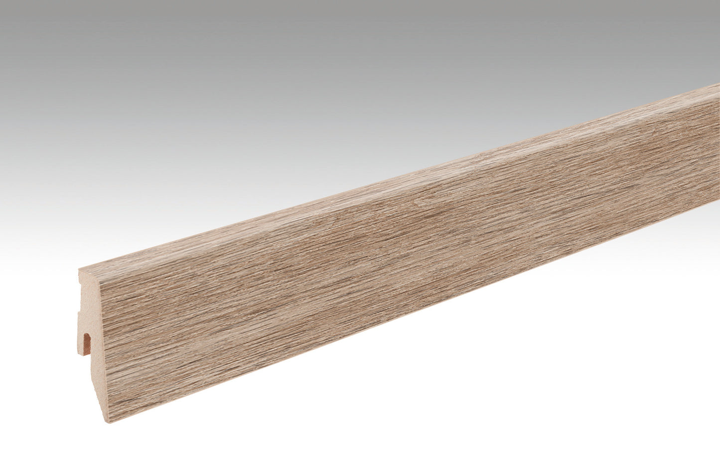 Eine lange, schmale Fußleiste mit holzähnlicher Maserung. Die Leiste ist leicht abgeschrägt und hat eine karamellfarbene Eichenoptik. Sie steht vor einem neutralen, hellgrauen Hintergrund, sodass ihre strukturierte Oberfläche und die Kanten deutlich erkennbar sind. Die Unterseite der Fußleiste zeigt eine kleine Aussparung zur Befestigung.