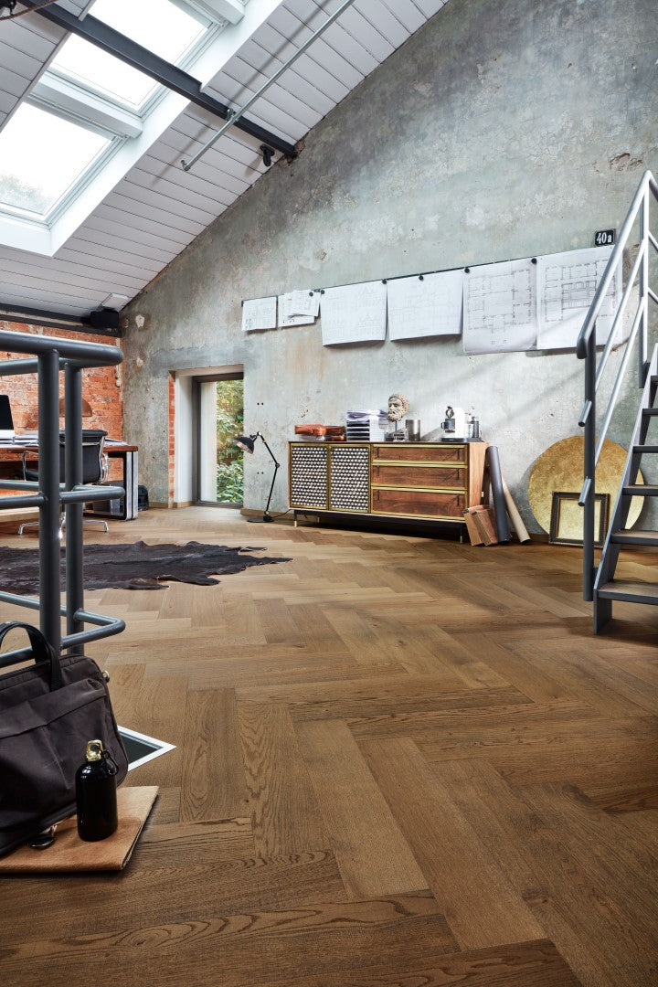 Großer Raum mit modernem Industrie-Look, Fischgrät-Holzboden, große Fenster, sichtbare Backstein- und Putzwand, Schreibtisch mit Stuhl, Kommode mit Büroartikeln, schwarze Stehlampe, schwarze Fell-Teppich, Tasche und Flasche am Boden, Metalltreppe, an der Wand hängen Zeichnungen oder Pläne.