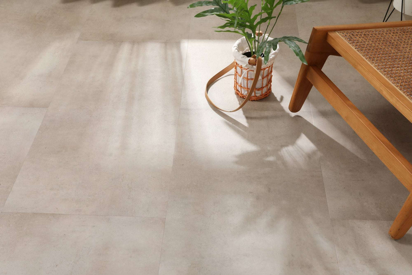 Auf dem Bild sind große, helle Fliesen zu sehen. Darauf steht ein Korb mit einem grünen Blattpflanze. Rechts unten ist ein Holzmöbelstück mit geflochtener Sitzfläche zu erkennen. Sonnenlicht wirft Schatten auf den Boden.