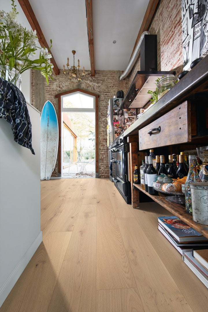 Heller Parkettboden mit Holzoptik, Surfbrett an der Wand, Glastür zum Garten, offenes Küchenregal mit Weinflaschen, Blumen, Bücher, freiliegende Backsteinwand, Deckenbalken, Kronleuchter, Küchenutensilien.