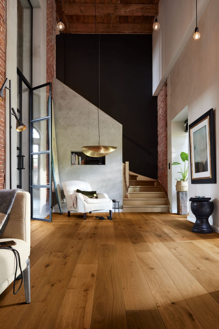 Ein Wohnzimmer mit Parkettboden aus Eiche, eine cremefarbene Polstersessel, kleiner Beistelltisch, goldfarbene Hängelampe, moderne Treppe, Wand mit Ziegel und Betonoptik, hohe Decke mit sichtbaren Holzbalken, Glastür, Zimmerpflanze in hellem Topf, Wandbild, schwarze Vase, Regal mit Büchern, moderne Beleuchtung mit Glühbirnen.