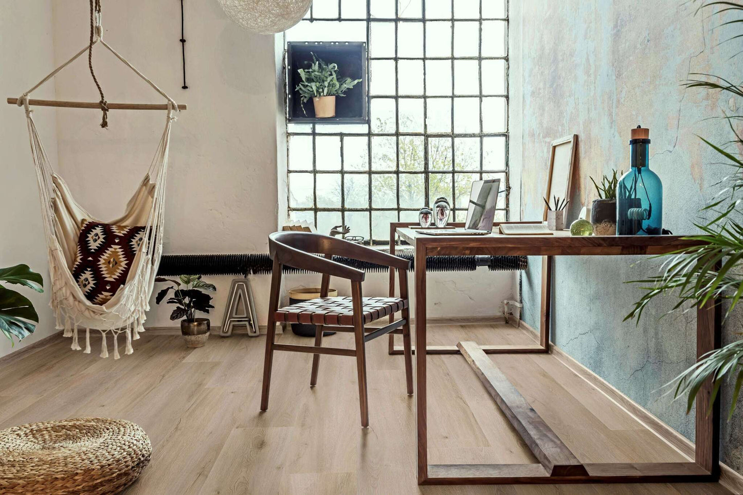 Ein heller Raum mit einem großen Fenster aus Glasbausteinen, einem Holztisch mit Laptop, Büchern, Pflanzen, blauer Flasche, Holzstuhl mit Lederbespannung, Hängesessel mit Kissen im Ethnomuster, Teppich aus Naturfasern, mehreren Zimmerpflanzen, Wanddekoration, Buchstabenobjekt "A"