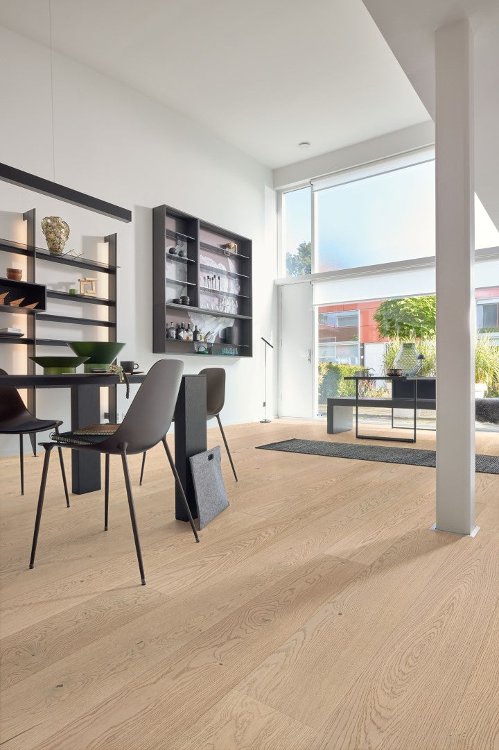 Heller Raum mit großen Fenstern, Holzboden, modernem Esstisch, zwei Stühlen, Wandregalen mit Deko, Sideboard, Objekte wie Schalen, Vasen, Lampe, Blick nach draußen auf Garten und rote Häuser, aufgeräumte und minimalistische Einrichtung.