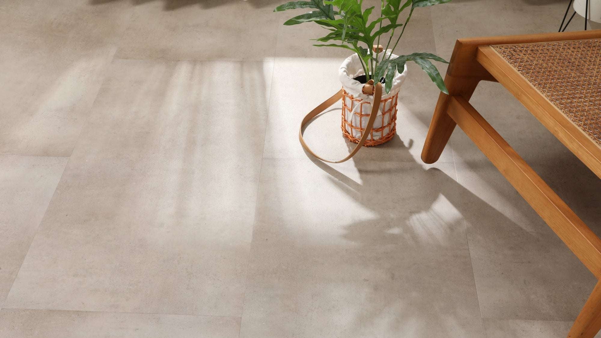 Auf dem Bild sind große, helle Fliesen zu sehen. Darauf steht ein Korb mit einem grünen Blattpflanze. Rechts unten ist ein Holzmöbelstück mit geflochtener Sitzfläche zu erkennen. Sonnenlicht wirft Schatten auf den Boden.