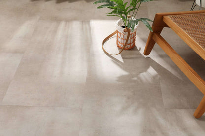 Auf dem Bild sind große, helle Fliesen zu sehen. Darauf steht ein Korb mit einem grünen Blattpflanze. Rechts unten ist ein Holzmöbelstück mit geflochtener Sitzfläche zu erkennen. Sonnenlicht wirft Schatten auf den Boden.