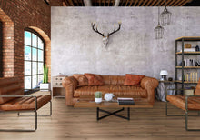 Großes Wohnzimmer mit Backsteinwand und großem Fenster, braunes Ledersofa und zwei Sessel, Couchtisch mit Kaktus, Teekanne, Buch, Regale mit Deko, Geweih an Betonwand, zwei hängende Lampen, Pflanzen und verschiedene Dekorationsobjekte.