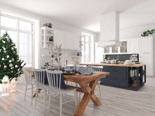 Heller Küchenraum mit großem Holz-Esstisch, sechs weißen Stühlen, festlicher gedeckter Tafel, geschmücktem Weihnachtsbaum, Kücheninsel, weißen Schränken, Pflanzen und Dekorationen.