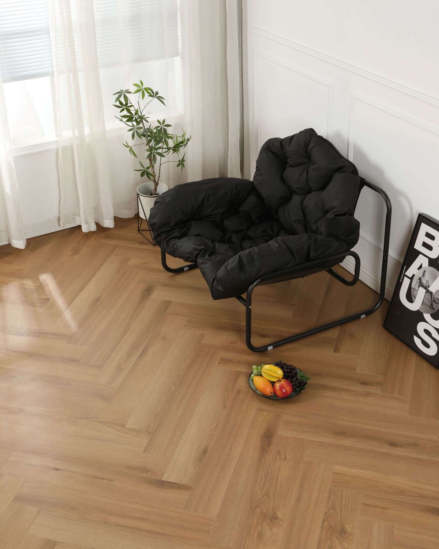 Schwarzer gepolsterter Sessel mit Metallrahmen, daneben kleine grüne Zimmerpflanze im weißen Topf, Fruchtschale mit Trauben, Banane, Mango und Gemüse auf hellem Holzparkett, neben dem Sessel ein gerahmtes Poster an der weißen Wand, Fenster mit weißen Vorhängen im Hintergrund.