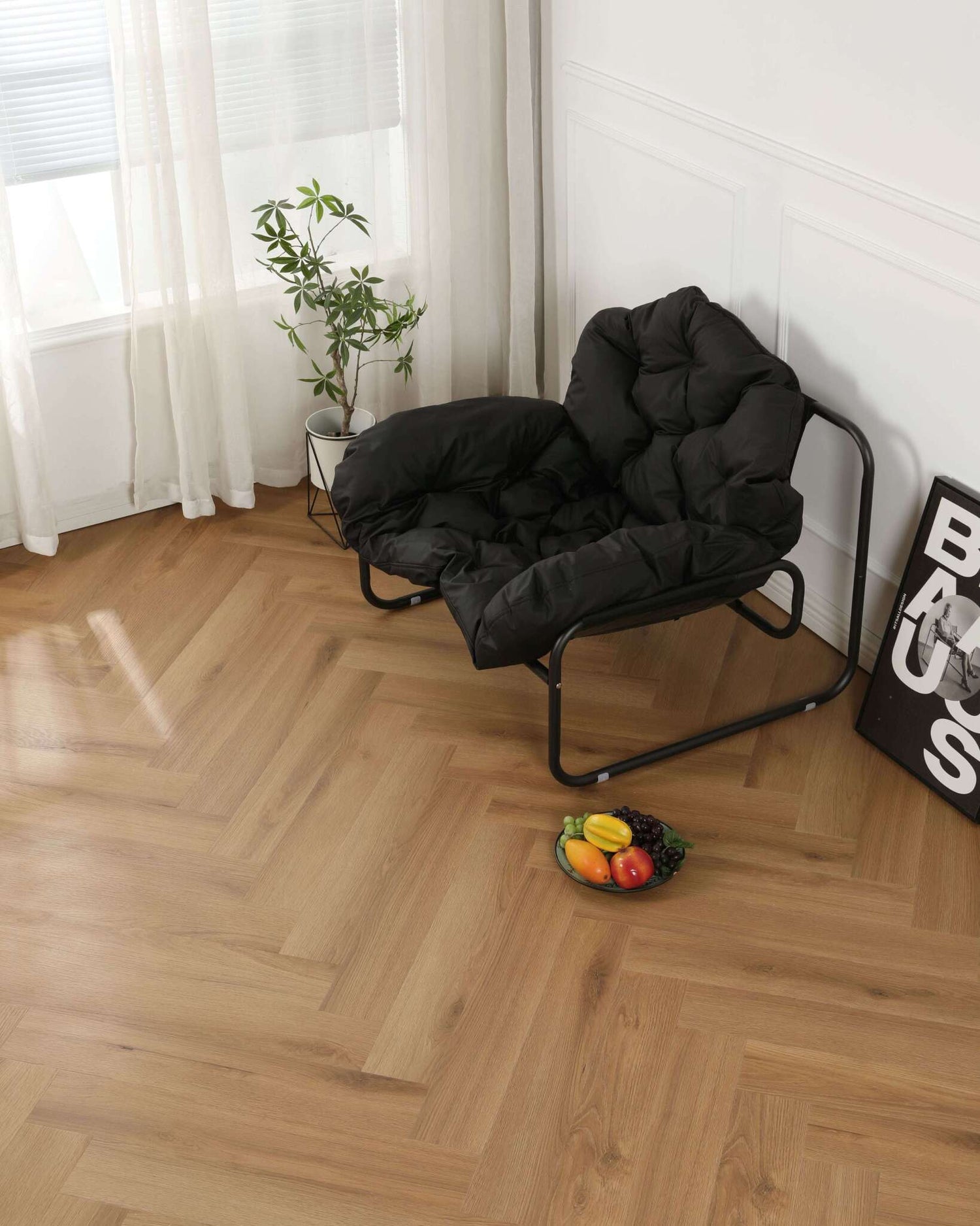 Schwarzer gepolsterter Sessel mit Metallrahmen, daneben kleine grüne Zimmerpflanze im weißen Topf, Fruchtschale mit Trauben, Banane, Mango und Gemüse auf hellem Holzparkett, neben dem Sessel ein gerahmtes Poster an der weißen Wand, Fenster mit weißen Vorhängen im Hintergrund.