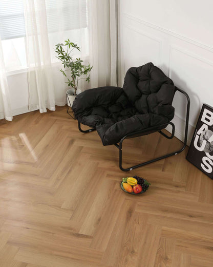 Schwarzer gepolsterter Sessel mit Metallrahmen, daneben kleine grüne Zimmerpflanze im weißen Topf, Fruchtschale mit Trauben, Banane, Mango und Gemüse auf hellem Holzparkett, neben dem Sessel ein gerahmtes Poster an der weißen Wand, Fenster mit weißen Vorhängen im Hintergrund.