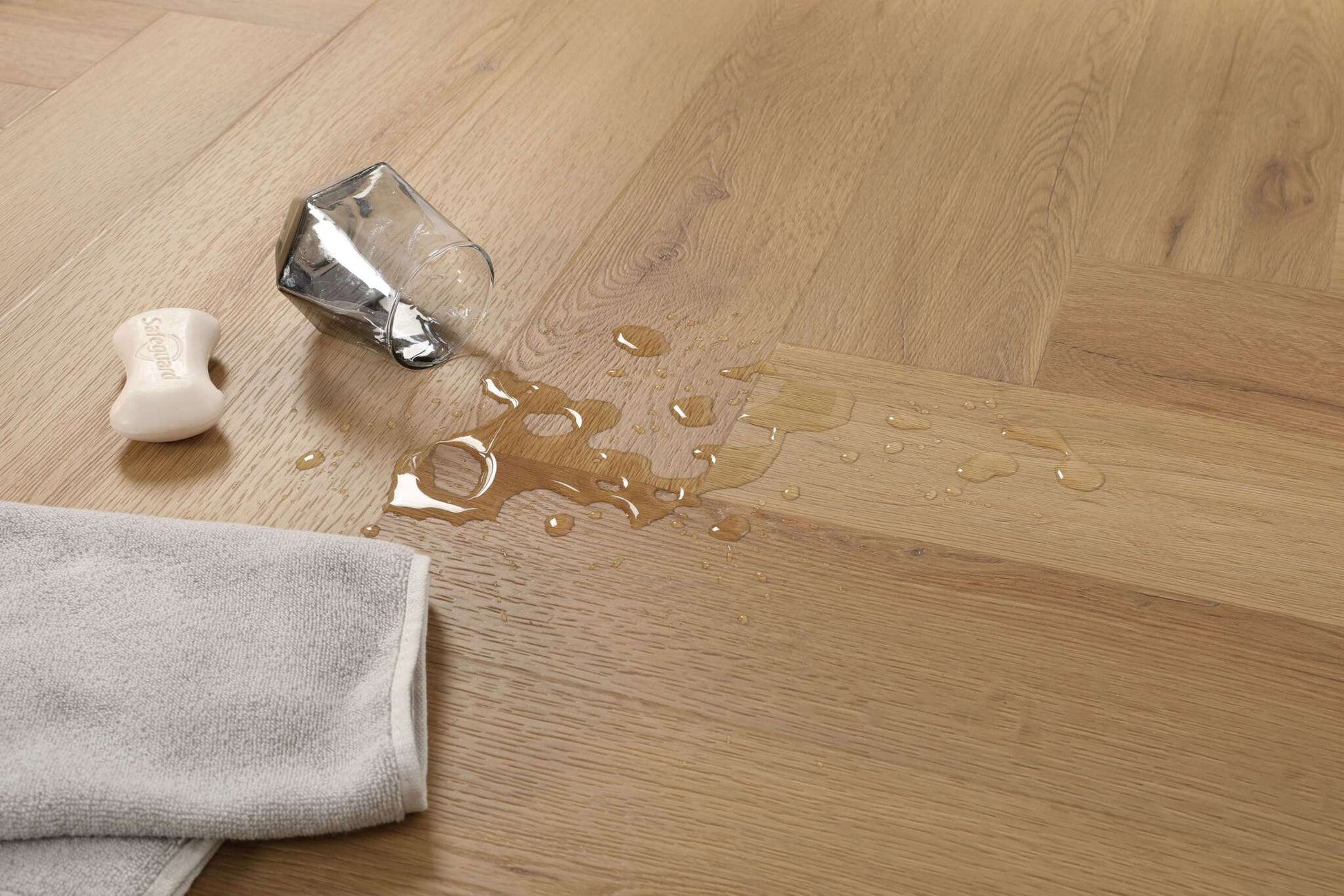 Auf einem Holzfußboden liegen ein umgekipptes Glas mit Wasser- oder Flüssigkeitsflecken, ein Stück Seife und ein graues Handtuch.