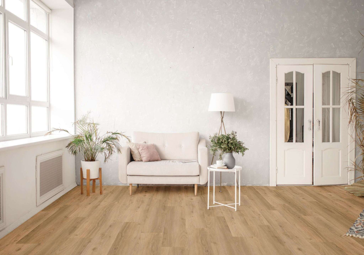 Ein helles Wohnzimmer mit beigen Sofa, zwei Kissen, weißem Beistelltisch, grüner Pflanze, Stehlampe, weißen Doppeltüren, großen Fenstern und Holzboden. Links eine Topfpflanze auf Holzständer, rechts weitere Pflanzen