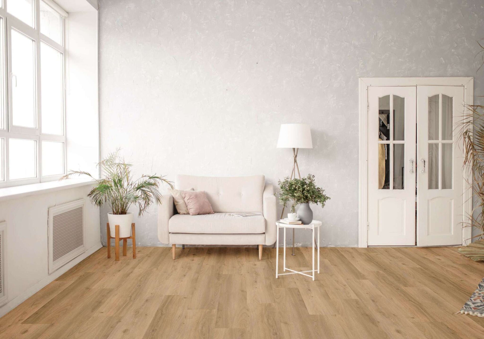 Ein helles Wohnzimmer mit beigen Sofa, zwei Kissen, weißem Beistelltisch, grüner Pflanze, Stehlampe, weißen Doppeltüren, großen Fenstern und Holzboden. Links eine Topfpflanze auf Holzständer, rechts weitere Pflanzen