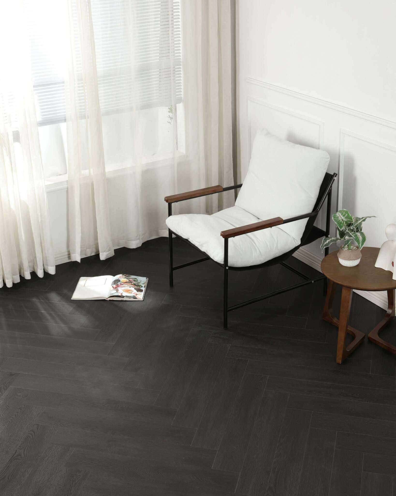 Ein moderner Sessel mit weißem Polster und Holzarmlehnen steht neben einem runden Holztisch mit Zimmerpflanze darauf. Ein offenes Magazin liegt auf dem dunklen Fischgrät-Parkettboden vor dem Sessel. Im Hintergrund helle Vorhänge vor Fenster und weiße Wandverkleidung.