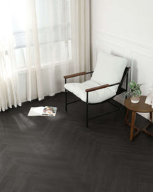 Ein moderner Sessel mit weißem Polster und Holzarmlehnen steht neben einem runden Holztisch mit Zimmerpflanze darauf. Ein offenes Magazin liegt auf dem dunklen Fischgrät-Parkettboden vor dem Sessel. Im Hintergrund helle Vorhänge vor Fenster und weiße Wandverkleidung.