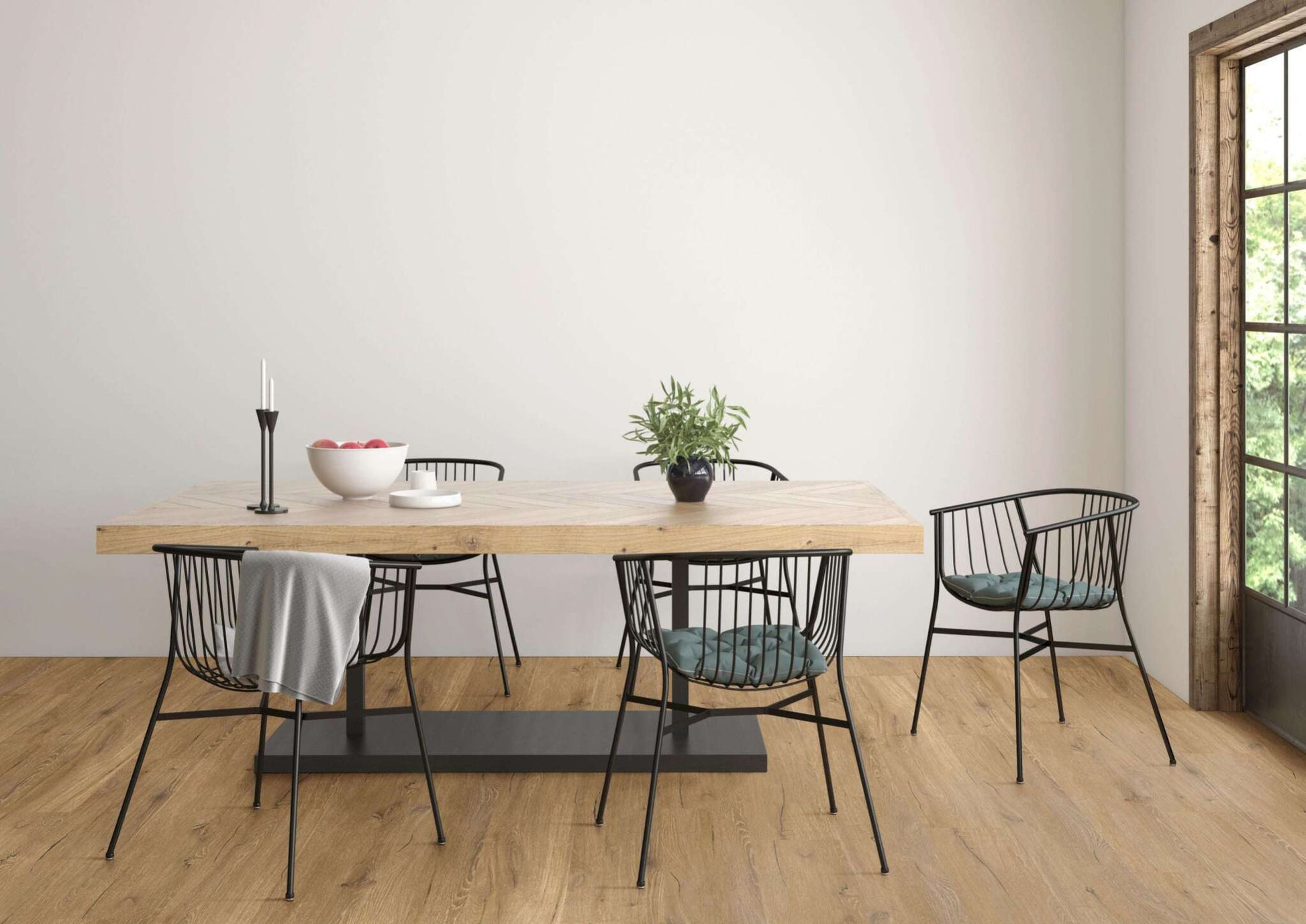 Holztisch mit schwarzem Metallgestell, fünf schwarze Stühle mit Drahtgestell, graue Decke, grüne Sitzkissen, kleine Pflanze, weiße Schale mit Äpfeln, schwarze Kerzenhalter mit weißen Kerzen, Fensterrahmen aus Holz, heller Raum, Holzboden.