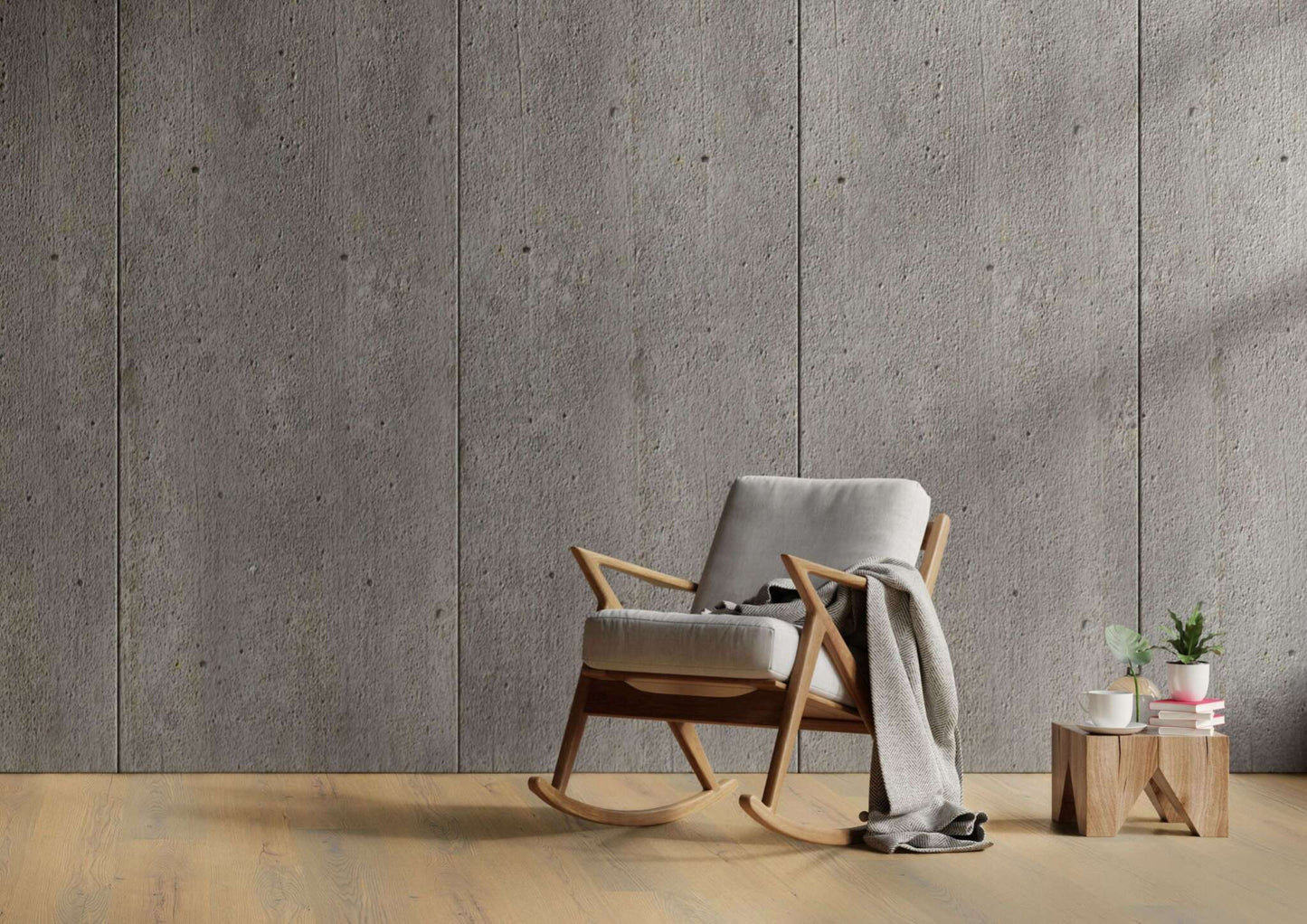 Ein moderner Schaukelstuhl mit hellgrauer Polsterung und Holzrahmen, darauf eine graue Decke. Daneben niedriger Holztisch mit zwei Pflanzen in weißen Töpfen, einer weißen Tasse, einer Untertasse und gestapelten Büchern auf hellem Holzboden, Hintergrund Betonwand.