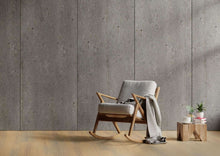 Ein moderner Schaukelstuhl mit hellgrauer Polsterung und Holzrahmen, darauf eine graue Decke. Daneben niedriger Holztisch mit zwei Pflanzen in weißen Töpfen, einer weißen Tasse, einer Untertasse und gestapelten Büchern auf hellem Holzboden, Hintergrund Betonwand.